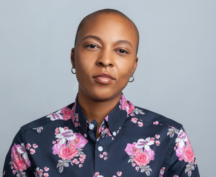 TJ is a non-binary person with lighter skin and a shaved head. They are wearing a blue button-down shirt with pink flowers. They have on small silver hoop earrings.